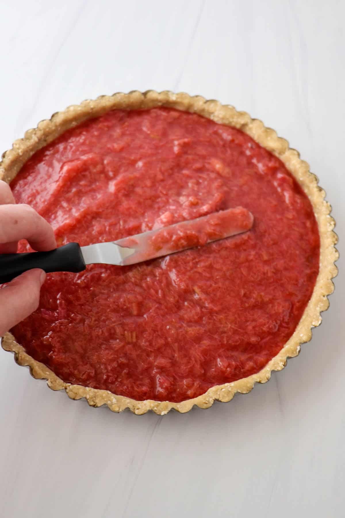 Tart crust topped with rhubarb filling being smoothed by an offset spatula.