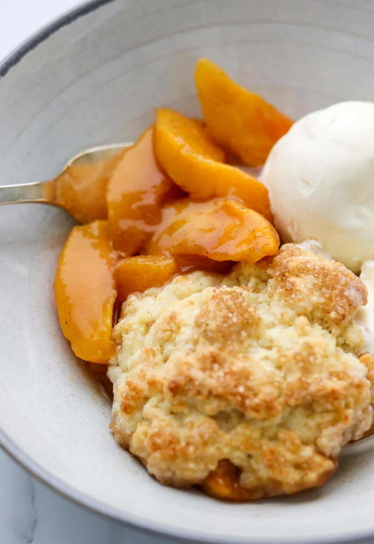 Serving of Cardamom Peach Cobbler in a bowl with ice cream and a fork.
