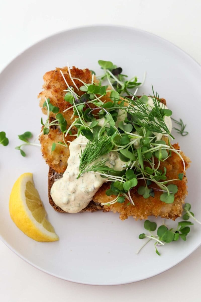 Pan-fried fish on rye bread topped with sauce, greens and with a lemon wedge.