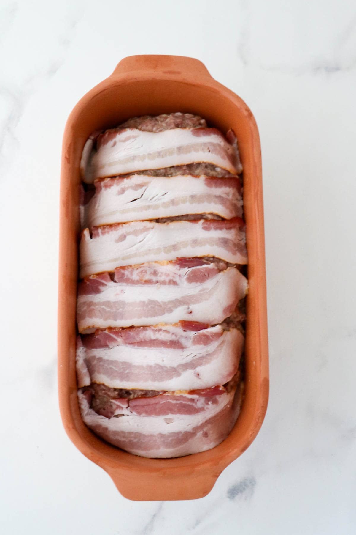 Danish meatloaf in a clay baker topped with bacon slices.
