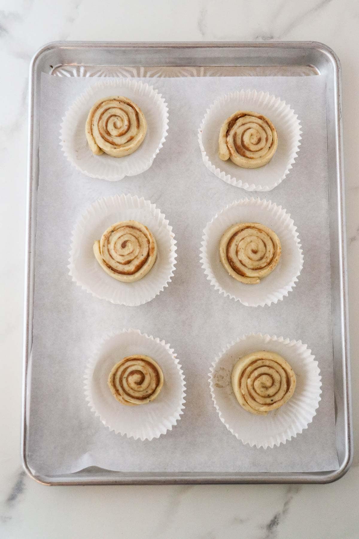 Six unbaked Norwegian Cinnamon Buns (Skillingsboller) on a baking sheet.