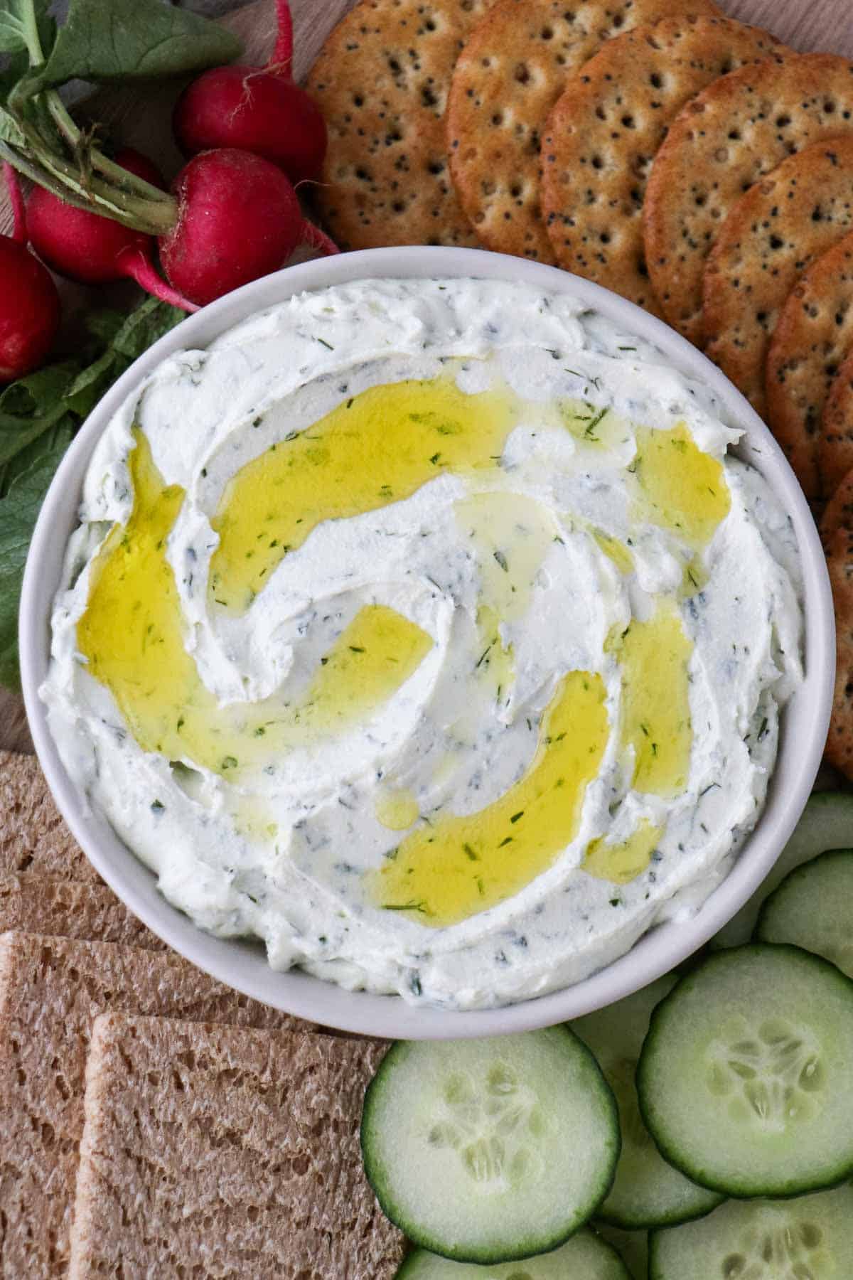 Bowl of Easy Herbed Whipped Goat Cheese in a bowl drizzled with olive oil and surrounded by crackers, cucumbers and radishes.