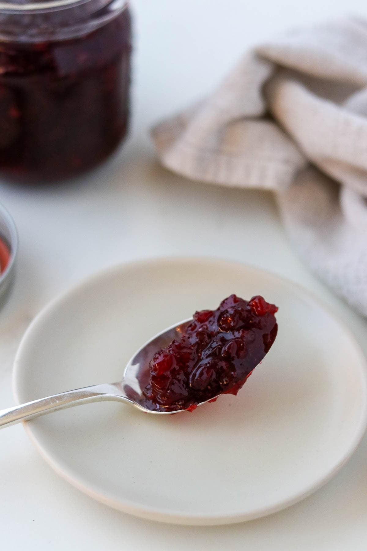 Homemade Lingonberry Jam on a spoon sitting on a white plate next to a tea towel and a jar of jam.