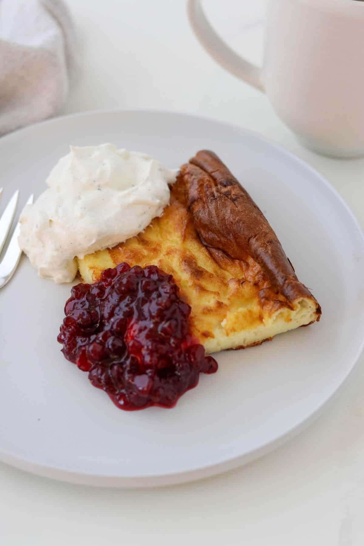 Slice of Swedish Oven Pancake (Ugnspannkaka) on a white plate with whipped cream and lingonberries.