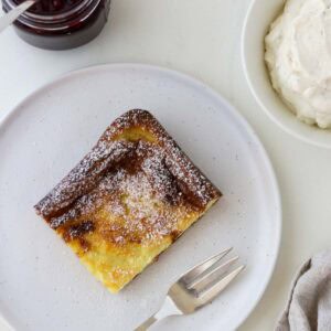 Slice of Swedish Oven Pancake (Ugnspannkaka) on a white plate with a fork, whipped cream and lingonberry jam.