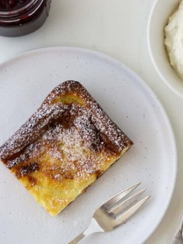 Slice of Swedish Oven Pancake (Ugnspannkaka) on a white plate with a fork, whipped cream and lingonberry jam.